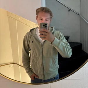 Ljusgrön hoodie från Polo Ralph Lauren - Säljer en ljusgrön hoodie från Polo Ralph Lauren i storlek M. Tröjan har dragkedja, huva med snörning och två fickor framtill. Klassisk broderad logga på bröstet. Perfekt för chill dagar och enkel att matcha med jeans eller shorts. Har tappat metall saken på ett av snörena 