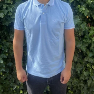 Ljusblå Fred Perry piké - Snygg ljusblå pikétröja från Fred Perry med klassisk krage och två knappar framtill. Diskret logga broderad på bröstet och vita ränder på krage och ärmslut. Tillverkad i mjuk bomull som känns skön mot huden. Perfekt för dig som gillar stilrena och sportiga plagg. .     Nypris: 1099kr GORILLAPRIZE:649kr             Skick:10/10 oöppnad          KVITTO MEDKOMMER 