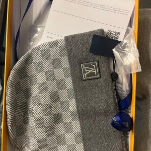 Grå rutig mössa Louis Vuitton - Exklusiv grå mössa från Louis Vuitton med rutigt mönster och stilren LV-logga framtill. Mössan har bred ribbad kant och är tillverkad i mjukt material som känns lyxigt mot huden. Perfekt accessoar för dig som vill ha en snygg och trendig look.