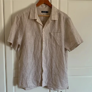 Beige linneskjorta från J.Lindeberg - Snygg beige skjorta från J.Lindeberg i svalt linne. Skjortan har korta ärmar, klassisk krage, bröstficka och knäppning framtill. Pris kan diskuteras