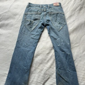 True Religion ljusblå jeans, stl 30 - Säljer ett par ljusblå jeans från True Religion med klassiska stora bakfickor och tydliga kontrastsömmar. Jeansen har slitningar och hål på både fram- och baksida för en cool, avslappnad look. Har Liten passform.