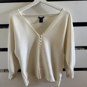 Beige kofta med knappar från Lindex - Superfin beige kofta från Lindex med v-ringning och snygga pärlemorknappar framtill. Koftan har en mjuk känsla, trekvartsärmar och en clean, stilren look som passar perfekt till jeans eller kjol.