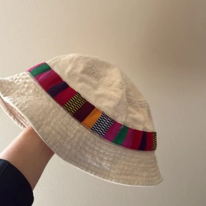 Vit bucket hat med färgglad rand från Pull & Bear - Bucket hat i vit tyg från Pull & Bear med en bred, färgglad rand runt hatten. Randen har inslag av grönt, rosa, lila, gult, orange och svart i ett randigt mönster. Hatten ser väldigt använd ut, men har använt ett fåtal gånger och är i fint skick annars. Storlek M. 