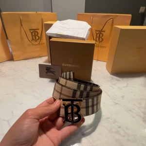 Burberry rutigt bälte med TB-spänne - Snyggt Burberry-bälte med klassiskt rutig design i beige, svart och brunt. Bältet har ett stort metallspänne med Burberrys ikoniska TB-logo. Tillverkat i skinn och har exklusiv känsla, perfekt för att lyfta din outfit med en lyxig touch.