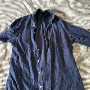 Blå linneskjorta Polo Ralph Lauren - Snygg mörkblå skjorta från Polo Ralph Lauren i linne med klassisk krage och vita knappar. Slim fit-modell med lång ärm och ikonisk röd logga broderad på bröstet. Perfekt för dig som gillar stilrena och bekväma plagg.