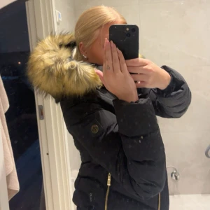 Svart pufferjacka Hollies med päls - Säljer en svart pufferjacka från Hollies med snygga guldiga dragkedjor och stor huva med fluffig päls. Insidan har leopardmönster som ger extra edge. Jackan är långärmad och perfekt för kalla dagar. Den är storlek 32 (xs) jag säljer den då den blivit för liten och bara ligger i garderoben. Köpt för 3år sen men bara legat i 2år. Den är knappt använd och är i nyskick. 