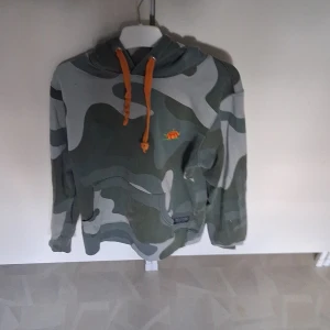 Camouflage hoodie från SQRTN Company - Säljer en grå och mörkgrön camouflage hoodie från SQRTN Company. Tröjan har orange snören i huvan Perfekt för dig som gillar streetwear och vill sticka ut lite extra.