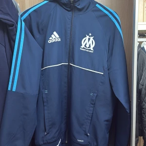 Marseille Marinblå vindjacka OM x Adidas  - Säljer en snygg marinblå vindjacka från Adidas i samarbete med Olympique de Marseille. Jackan har huva, dragkedja, vita detaljer och ljusblå ränder längs ärmarna. Perfekt för dig som gillar fotboll och vill ha en sportig look. Climalite-material. Riktig bra skick