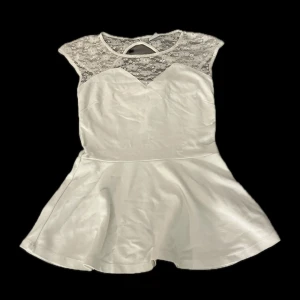 Vit tröja med volanger och spetsdetaljer - Supersöt vit topp med volanger som en ”tjol” ned till. med rund hals och spets upptill som ger en romantisk vibe. Tröjan har en ärmlös design och ett lätt utsvängt kjolparti. Spetsen har blommigt mönster och en snygg öppning i ryggen. Axelbredd-45cm. Längd:64cm.