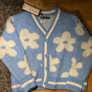 Blå fluffig kofta med blommor från FJ - Supermysig blå kofta med stora vita blommor och ribbade muddar. Koftan har v-ringad hals,  Materialet är fluffigt och stickat, perfekt för vinter , den är helt ny, säljs eftersom jag inte vet hur jag ska styla den men vet att någon annan hade kunnat Stayla den SÅ BRA