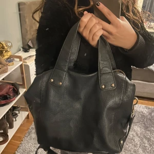Svart handväska från Anna Field - Snygg svart handväska från Anna Field i skinnimitation med guldfärgade detaljer och dragkedja. Väskan har både handtag och axelrem samt ett blommigt foder på insidan. Perfekt storlek för att få plats med allt du behöver till vardagen. Skavanker finns på sista bilderna 