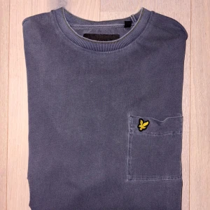 Grå t-shirt med ficka från Lyle & Scott - Snygg grå t-shirt från Lyle & Scott med klassisk rund hals och korta ärmar. På bröstet finns en ficka med den ikoniska gula fågel-loggan. T-shirten är gjord i mjuk bomull och har en relaxed passform som är enkel att matcha. Använd få gånger, modellen ska se sliten ut!