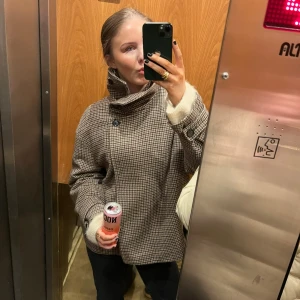 Rutig brun kappa med hög krage - Säljer en snygg rutig kappa i bruna och beige toner med hög krage och stora knappar. Jackan har lång ärm och en loose passform, perfekt för kyliga dagar. Klassisk och trendig look som passar till det mesta.