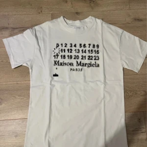 Maison Margiela vit t-shirt med tryck - Vit t-shirt från Maison Margiela med svart pixelgrafiskt tryck och logga på bröstet. Klassisk rund hals och korta ärmar. Materialet är mjuk bomull och passformen är normal. Perfekt för dig som gillar designerplagg med unik twist.