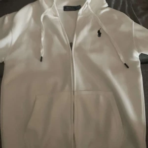 Vit hoodie från Polo Ralph Lauren - Vit hoodie från Polo Ralph Lauren med klassisk svart logga broderad på bröstet. Tröjan har huva med snörning, dragkedja framtill och en stor känguruficka. Materialet är mjuk bomull och passformen är normal. Perfekt för chill dagar.