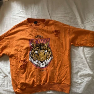 Orange sweatshirt med tigertryck Gina Tricot - Cool orange sweatshirt från Gina Tricot med stort tigertryck och texten 'Bronx New York' framtill. Tröjan har vita ränder på ärmarna och slitna detaljer. Mjuk bomullskänsla och avslappnad passform. Obs! Hålen är design
