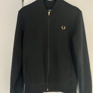 Svart jacka/tröja från Fred Perry - Svart jacka/tröja från Fred Perry med guldfärgad logga på bröstet och dragkedja framtill. Jackan har ribbade muddar, två fickor och klassisk bomberkrage. Perfekt för dig som gillar stilren och sportig look.