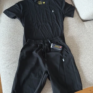 Svart t-shirt och shorts från Polo Ralph Lauren - Svart t-shirt från Polo Ralph Lauren med klassisk passform och rund hals. T-shirten har korta ärmar och en liten vit broderad logga på bröstet. Tillverkad i mjuk bomull som känns skön mot huden. Perfekt för en clean och stilren look.