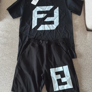 Svart Fendi t-shirt och shorts med stor logga - Svart t-shirt från Fendi med en stor vit Fendi-logga tryckt på bröstet. Klassisk rund hals och korta ärmar. T-shirten har en clean och modern look, perfekt för dig som gillar streetwear och vill sticka ut med ett lyxigt märke. Materialet är mjuk bomull.