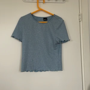 Glittrig blå topp från Gina Tricot - Supersöt glittrig blå topp från Gina Tricot i storlek M. T-shirten har korta ärmar och vågiga kanter vid ärmslut och nederkant. Perfekt för att lyfta vilken outfit som helst med lite extra shimmer.