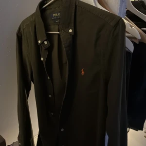 Grön  skjorta Polo Ralph Lauren slim fit - Mörkgrön långärmad skjorta från Polo Ralph Lauren i slim fit-modell. Klassisk design med knappar framtill och broderad logga i orange på bröstet. Skjortan är tillverkad i Manchester  och har en stilren krage. Perfekt för dig som gillar en clean och tidlös look.