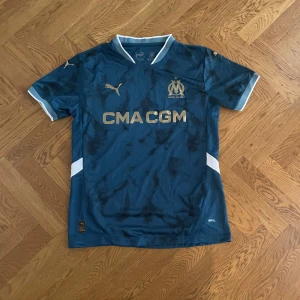 Marinblå Marseille fotbollströja Puma - Snygg marinblå Olympique de Marseille fotbollströja från Puma med guldfärgade detaljer och loggor. Kortärmad modell med vit kant vid krage och ärmslut, samt vita inslag på sidorna. Tillverkad i lätt och ventilerande material, perfekt för match eller träning.