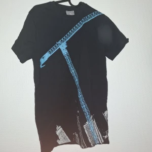 Svart t-shirt med blått tryck Fristads - Svart t-shirt från Fristads Kansas i storlek XS med coolt blått tryck av en byggkran och stadssilhuett på framsidan. Klassisk rund hals och korta ärmar. Passformen är normal och materialet känns som mjuk bomull.