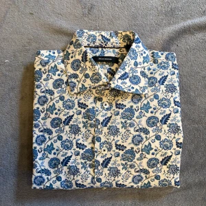 Matíníque skjorta - Vit och blå blommig finskjorta från Matíníque. Nyskick, aldrig använd. Hör av dig vid frågor, mått och bilder.                                                           • Storlek M/40 Herr.                                               • Längd 74cm.                                                           • Armhåla till Armhåla 51cm                                  • Axelbredd 42cm