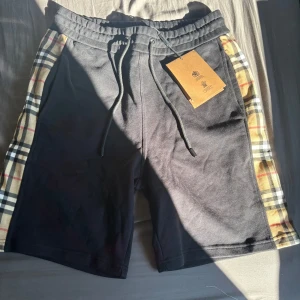 Svarta Burberry shorts  - Svarta shorts från Burberry med klassiska rutiga paneler längs sidorna i beige, svart och rött. Mjuk bomullskvalitet, elastisk midja med snörning och bakficka. Perfekt för en chill och stilren look.