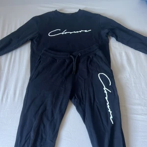 Svart tracksuit långärmad tröja/Byxa Closure London - En Svart tracksuit, byxa/ långärmad tröja från Closure London med vit logotyp framtill. Tröjan har rund hals, ribbade muddar och är gjord i mjuk bomull. Perfekt för chill dagar eller när du vill ha en clean streetstyle-look.