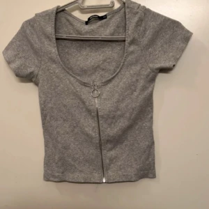 Grå ribbad topp med dragkedja Bik Bok - Snygg grå ribbad topp från Bik Bok i storlek XS. Modellen är croppad med korta ärmar och har en cool silverfärgad dragkedja framtill med ringdetalj. Perfekt för dig som gillar en enkel men trendig look.