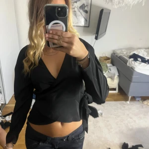 Svart omlottopp från Gina Tricot - Snygg svart omlottopp från Gina Tricot med långa ärmar och knyt i sidan. Djup v-ringning och croppad passform som ger en trendig look. Perfekt att styla med jeans eller kjol för en cool outfit.