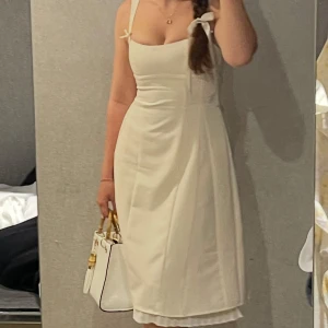 Oh polly klänning med rosetter - Supersöt vit klänning med omlott-detalj och rosetter på axelbanden. Klänningen har en figurnära passform upptill och lätt utsvängd kjol, vilket ger en feminin siluett. Axelbandslös känsla med smala band och snygg v-ringning. Perfekt för sommarkvällar!