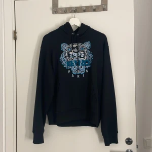 Svart Kenzo hoodie med tigerbrodyr - Svart hoodie från Kenzo i 100% bomull med ikonisk broderad tiger i blått och vitt på bröstet. Klassisk huva med snörning och känguruficka framtill. Perfekt för dig som vill sticka ut med en streetstyle-look.