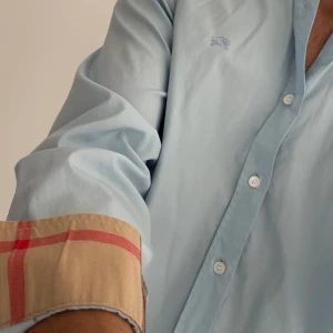 Ljusblå Burberry skjorta  - Snygg ljusblå skjorta från Burberry med klassisk rutig insida på krage och manschetter. Skjortan har knappar hela vägen, lång ärm och en liten broderad Burberry-logga på bröstet. Perfekt för dig som gillar stilrena plagg med en twist. Passar medium perfekt