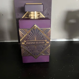 Infini Elixir parfym Maison Alhambra - Lyxig parfymflaska i lila med guldiga detaljer och geometriskt mönster. Flaskan har en fyrkantig form och en dekorativ kork. Märkt med Infini Elixir och Maison Alhambra på etiketten. Perfekt för dig som gillar exklusiva dofter.