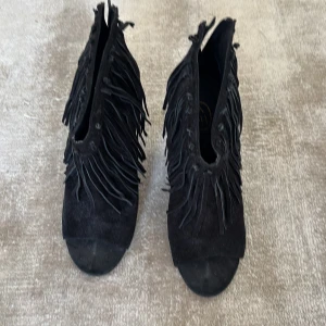Svarta mocka boots med fransar från ASH - Unika svarta boots i mocka från ASH med coola fransdetaljer runt hela skon. Modellen har öppen tå och en kilklack som ger extra höjd. Perfekta för dig som vill sticka ut med en edgy och trendig stil.