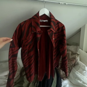 Röd zebramönstrad overshirt från Zara - Snygg röd overshirt från Zara med svarta zebraränder. Jackan har klassisk krage, två bröstfickor med lock och knappar framtill. Perfekt för dig som vill sticka ut med ett djurmönster och cool vibe. Långärmad och i mjukt bomullstyg.