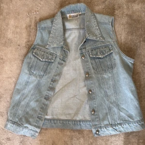 Ljusblå jeansväst med nitar - Snygg ljusblå jeansväst från Diamant med silvriga nitar på krage och bröstfickor. Klassisk jeansmodell med knappar framtill och två fickor på bröstet. Perfekt att slänga över en t-shirt för en cool look.