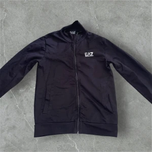 Svart bomberjacka från EA7 Emporio Armani - Svart bomberjacka från EA7 Emporio Armani med stilren design och diskret logga på bröstet. Jackan har dragkedja framtill, två fickor och ribbade muddar. Tillverkad i bomull och polyester, perfekt för dig som gillar en clean och sportig look.