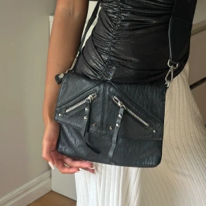 Svart axelväska med dragkedjor - Cool svart axelväska i skinn med två sneda dragkedjor och dekorativa nitar på framsidan. Väskan har bred axelrem och ett rymligt huvudfack med extra innerficka. Perfekt för att bära dina essentials och ge din outfit en edgy vibe.
