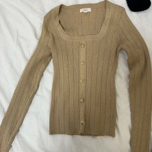 Beige ribbad kofta från Guaja - Snygg beige kofta från Guaja med ribbad struktur och guldfärgade knappar framtill. Tröjan har långärmad passform och en fyrkantig urringning som ger en modern vibe. Perfekt att styla med jeans eller kjol för en chill look.