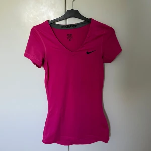 Nike Pro rosa t-shirt med v-ringning - Nike Pro t-shirt i rosa färg med svart logga på bröstet. V-ringad hals och korta ärmar. Slimmad passform.