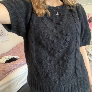 Svart stickad tröja & Other Stories - Mysig svart stickad tröja från & Other Stories med korta ärmar och chunky strukturstickning. Tröjan har ett coolt mönster med bollar och flätor framtill samt ribbade muddar. 