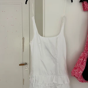 Vit kort klänning med volanger från Zara - Supersöt vit klänning från Zara med smala axelband och volangdetaljer nedtill. Klänningen har en rak, lite loose passform och är perfekt för varma dagar. Materialet känns lätt och fräscht, och den är ärmlös med en snygg, djup urringning framtill.