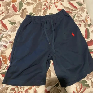 Mörkblå shorts från Polo Ralph Lauren - Mörkblå shorts från Polo Ralph Lauren med elastisk midja och snörning. Klassisk röd logga broderad på ena benet. Sköna och stilrena, perfekta för sommaren. Tillverkade i mjuk bomull för maximal komfort.