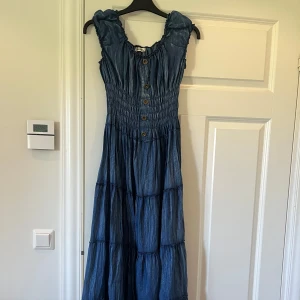 Blå långklänning med knappar - Supersöt blå långklänning i jeansimitation med smockad överdel, volangiga axelband och knappar framtill. Aldrig använd. 