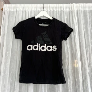 Svart Adidas t-shirt - Svart t-shirt från Adidas med stor vit och svart logga framtill. Klassisk rund hals och korta ärmar. Skön passform och tillverkad i mjuk bomull, perfekt för en sportig look.