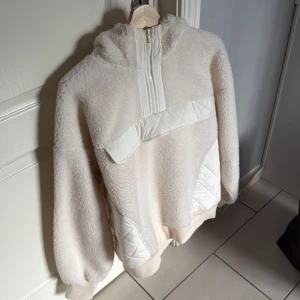 Beige fleecehoodie från H&M Move - Supermjuk och mysig beige hoodie från H&M Move i fleece med quiltade detaljer på axlar, fickor och bröst. Tröjan har huva, half zip och ribbade muddar. Perfekt för kyliga dagar när du vill vara både varm och snygg.