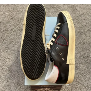 Philippe Model svarta sneakers i skinn - Svarta sneakers från Philippe Model i skinn med vit sula och vita snören. Skorna har en vit hälkappa med röd kant och en svart sköldpatch med röd kant på sidan. Snygg vintage-look på sulan och klassisk låg modell.